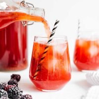 Pouring berry vanilla kombucha in a glass with a paper straw on white background
