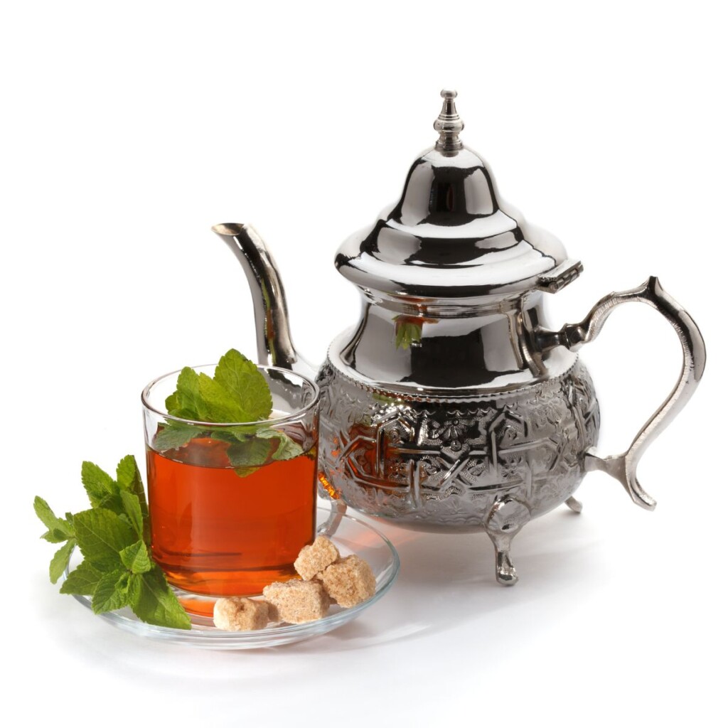 Moroccan mint tea in a cup next to a silver steeping pot on an isolated white background.
